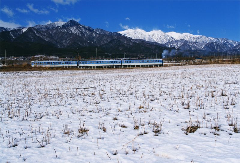雪が積もった田んぼの奥に電車が走っている写真です